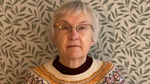 Contributed Inga Lockington is standing in front of a wall covered in patterned wall paper of green leaves. She is wearing a multi-coloured patterned jumper with a black roll neck underneath. It is a head shot and Inga is wearing glasses and looking straight at the camera. 