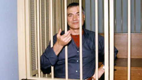 Mafia boss Nitto Santapaola during his trial for the assassinations of anti-mafia prosecutors. A man wearing a blue shirt is behind white bars and snaking his hand through the bars, with his other hand on his hips
