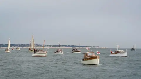 Multiple ships dating back from the 1940s sailing in the sea. Some of the ships are flying St George's flags. The English coast is sat in the background.