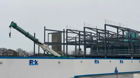 Une grue, à côté d'une structure en poutres d'acier, derrière des palissades bleues et blanches qui indiquent Rok Construction.