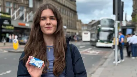 Poppy Fairbairn holds the National Entitlement Card towards the camera with a blurred Lothian Bus in the background.