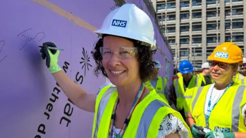 Doncaster and Bassetlaw Teaching Hospitals A woman with dark curly hair is wearing a white hard hat which says NHS. She has health and safety goggles and a high-vis vest and is smiling while signing her name on a purple hoarding on a building site
