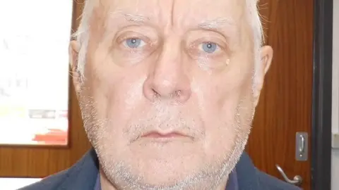 A man with blue eyes and thin, short grey hair stares expressionless into the camera. He has a stubbly beard and appears to be wearing a blue jumper.