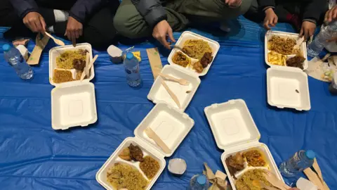 Boxes of food being shared while people are sat on a blue mat 