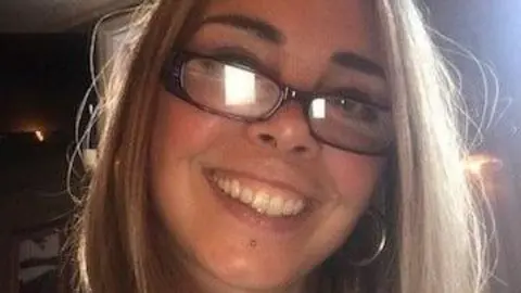 Delia McInerney, who has straight blonde hair, a piercing under her bottom lip and silver earrings. Wearing glasses, she smiles at the camera while taking a selfie. 