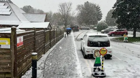 The Prince of Wales School Wooden fence, path and road with school road signs, several parked cars and snow falling 