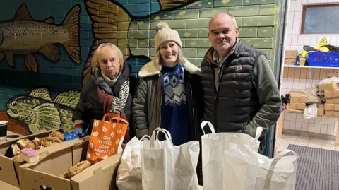 Maureen, Emma and Mark Briggs standing behind a table with bags of food and boxes of donated parties from local businesses.