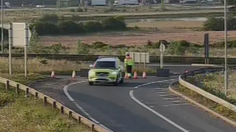 A marked fluorescent emergency vehicle on the lane of the A14 with cones in place.