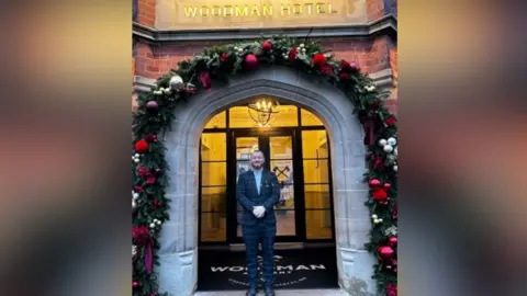 Ryan is stood in the doorway of the pub which has a large, stone arch withy floral decorations around it. A large black mat with the pub's name on can be seen behind him by glass doors at the entrance. Ryan is wearing a blue/green checked suit. he had red hair and a beard.