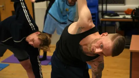 John Fairhall/BBC Adam Townsend is striking a yoga pose, with his arms outstretched and he is reaching down on one side. He is heavily tattooed, with a brown beard. He is wearing a black vest. Other members are doing the same pose in the background. 