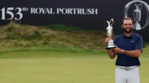 Reuters A man in a navy blue polo shirt, white trousers and a silver wrist watch holds up a trophy up and smiles at the camera. Behind him is a putting green and a sign which says "153rd Royal Portrush".