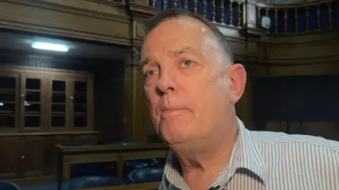 Andy Frampton looking to the left out of frame. He is wearing a striped button shirt and is standing in the council chamber, with wooden furniture and blue chairs in the background.