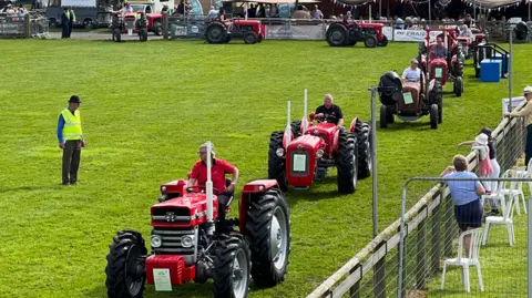 The image shows a number of tractors queuing up in a line in a showground. Most of them are red.