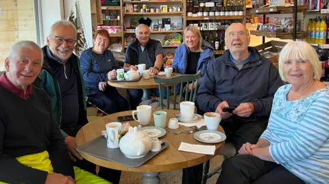 Seven people sit across two tables in a cafe, they have mugs and teapots in front of them. 