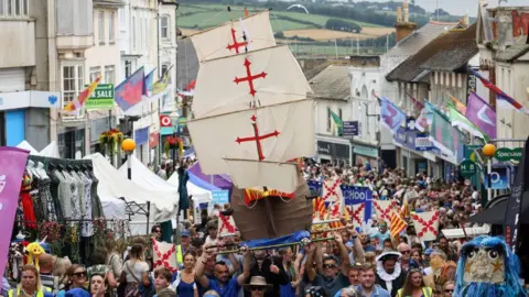 Penzance Council A street filled with people, with flags along the sides and a group of people holding a replica ship above the crowds