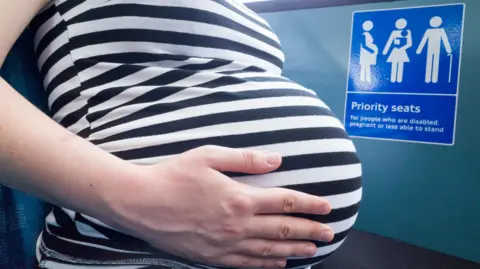 A close-up side profile of a pregnant woman's stomach in a striped shirt, standing next to a blue "Priority seats" sign on a train.
