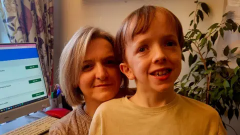 Wendy Gallagher and her son Andrew smiling at the camera. Andrew is slightly in front of Wendy. He has fair hair with a fringe and is wearing a yellow top. Wendy is wearing a beige jumper and has shoulder length fair hair. In the background, there is a computer and a green plant.