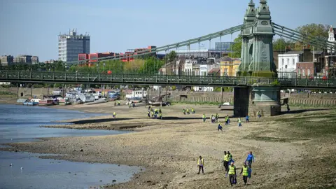 Getty Volunteers can be seen participating in a clean-up of a Thames riverbank. There are lots of people walking along a large stretch of beach under a green bridge wearing yellow hi-vis vests and carrying litter pickers and bin bags. 