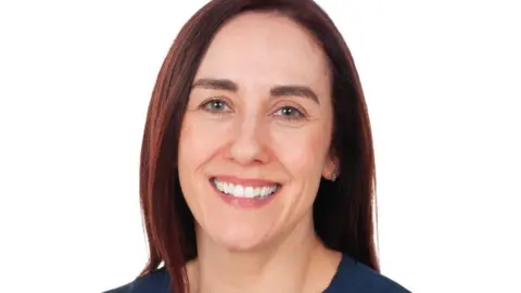 Bolton NHS Foundation Trust A person is seated against a plain white background, wearing a dark navy blue dress with short, wide sleeves and a fitted waist. The individual has straight, shoulder-length hair that is dark brown with a reddish tint. The posture is upright, with arms resting naturally in front.