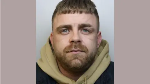 A police custody image of Warren Chiddy. He is wearing a green hoodie and has short hair and a reddish brown beard
