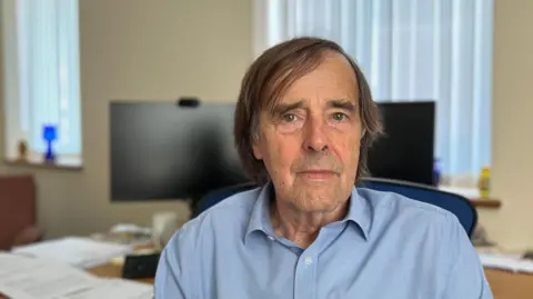 Constable Richard Honeycombe is sitting in his parish office, with two desktop screens behind him. He is wearing a blue shirt and is looking into the camera.