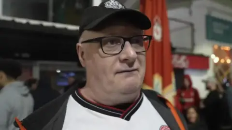 Paul Rodgers is wearing a white Chatham Town shirt, a hat and glasses. Behind him the background is blurred but we can see other people.