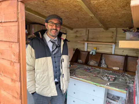 Steve stands inside a wooden shed wearing a winter jacket over formal clothing, beside a detailed model town set on top of cabinets. The shed’s interior is lined with wooden panels and shelves holding tools, boxes, and assorted materials.