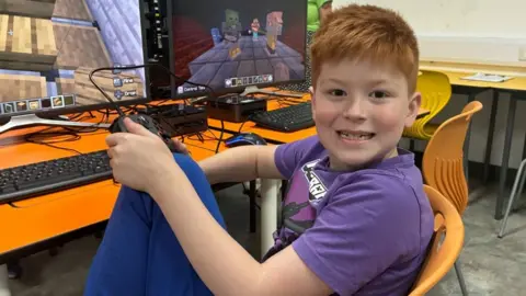 A young boy with short ginger hair and freckles wears a purple graphic t-shirt and blue joggers. He sits at a desk with a large monitor and holds a game console controller.