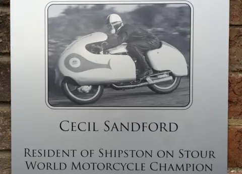 Ian Sandford A silver plaque with a picture of a man riding a motorbike. Beneath it says "Cecil Sandford resident of Shipston on Stour, World Motorcycle Champion"