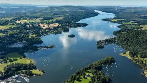 An aerial view of Windermere, England's largest lake which stretches into the distance narrowing as it goes. Boats can be seen dotted along its foreshore