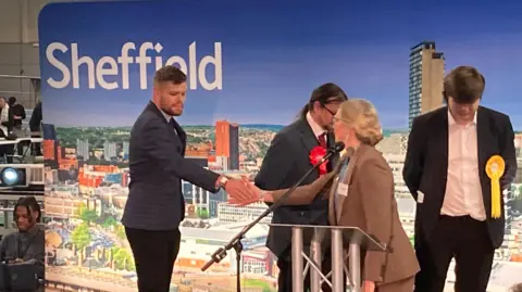 BBC/Simon Thake Candidate Nathaniel Menday shakes the hand of the Sheffield returning Officer after winning his election in Woodhouse. He is wearing a dark suit and has a thin beard. He is standing in front of a banner with the words Sheffield on it.