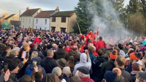 Jake Zuckerman/BBC A large crowd of people prepare to take part in a medieval rugby-style game in the centre of a village. They are surrounding the fool, who is dressed in red and holding a leather tube (the ball) aloft. Some people are filming the spectacle on their phones. Houses and trees can be seen in the background.