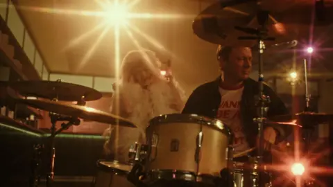 Eilís Doherty/AWAL Jamie Oliver playing the drums in a studio looking setting. There is a lot of lens flare from lights shining at the camera. A woman can be seen listening enthusiastically in the background wearing a white hooded top.