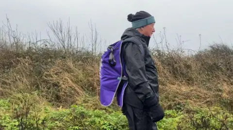 Jadzia Samuel/BBC Derrick Downs walking in a long shot from left to right with the top of a cliff and the sea behind him