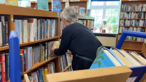 The photo shows a woman putting a book back on a book shelf. She's wearing a black cardigan, and has a black cross body bag too. There are also lots of shelves filled with books. 
