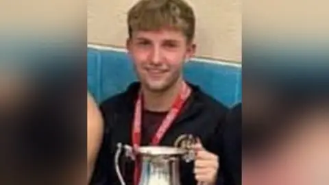 A picture of Cain Pears holding a football trophy and wearing a medal with a red ribbon around his neck. He has short dark blond hair and is smiling.