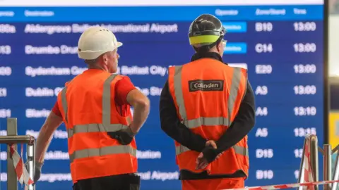 Pacemaker Two men wearing hi-vis vests and hard hats looking at a sign showing bus departures 