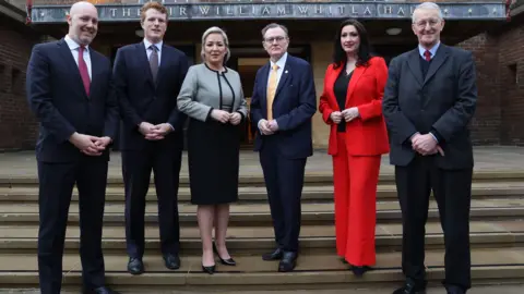 PA Media James Applegate, Joe Kennedy, Michelle O'Neill,Professor Sir Ian Greer,deputy Emma Little-Pengelly and Northern Ireland Secretary Hilary Benn, pose for a photograph on day three on the steps of Whitla Hall at Queen's University Belfast.