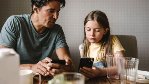 A stock image of a man and child looking at their phones at the breakfast table