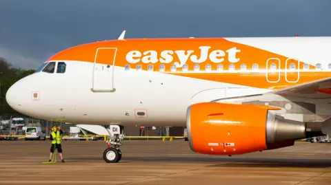 The front of a plane parked at an airport, branded with the word easyJet, with a streak of orange paint over the body of the plane. A person in a high-vis vest is standing under the plane.