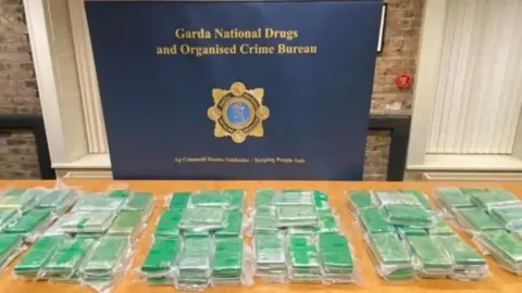 An Garda Síochána Six Green cocaine parcels, laid out on a wooden table with a Garda Natiional Drugs banner and badge displayed in background