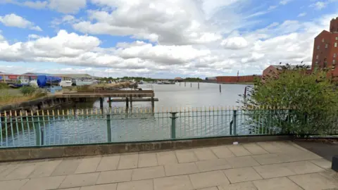 View from a bridge of the River Freshney in Grimsby where the body was discovered. There is a green and gold metal fence with wharves and industrial land in the background
