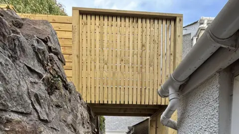 PEDW A timber raised platform connecting a bricked wall and a grey bricked house. 