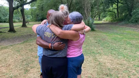 Four adults hug each other in a park. Three are women and there is one man.