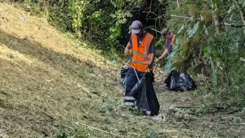Slough Borough Council/LDRS Men in orange high-visibility jackets clearing rubbish in a grassy area using litter pickers and bin bags