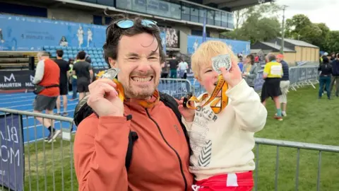 A man holds his son. Both are smiling and proudly holding up medal to the camera.