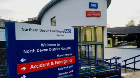 A sign which reads: Northern Devon Healthcare NHS Trust, Welcome to North Devon District Hospital, and there are arrows pointing to the Main Entrance, Accident and Emergency, Ladywell Unit and Seamoor Unit. There is a building behind the sign with a double door.