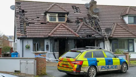 Pacemaker A police car is parked outside a house which has been gutted in a fire - it is a chalet-bungalow style and the front door and front window are completely gone, surrounded by charred frames. Inside the living room it is black with soot. Guttering has collapsed and debris lies on the paved driveway. 