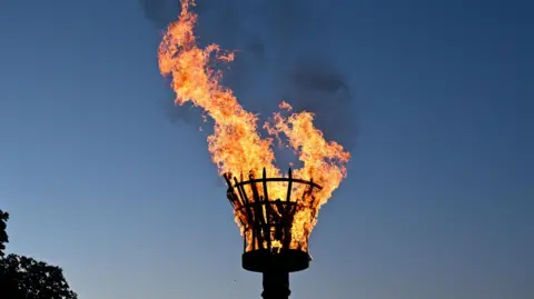A ceremonial beacon, formed out of a metal basket, on fire against a twilight sky