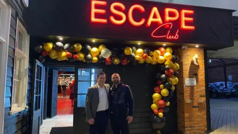 Ervin Hyso Steven Wells and Ervin Hyso standing in front of the Escape nightclub in Ipswich. Steven is wearing a blazer over a white shirt while Ervin is wearing a dark navy gilet. They are standing underneath an multi-coloured balloon arch. 